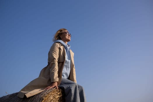 A woman is sitting on a hay bale, wearing a tan coat and a blue hoodie. She is smiling and looking up at the blue sky