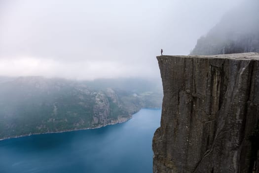 A lone figure stands on the edge of Preikestolen, a dramatic cliff in Norway, overlooking a vast expanse of water shrouded in mist. Preikestolen, Norway