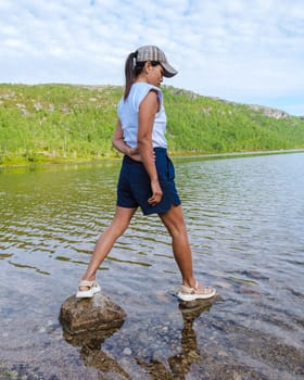 A woman wearing a white shirt and blue shorts walks across stones in a lake in Norway, with lush green hills in the background.