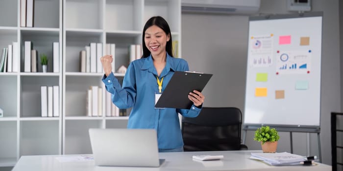 Successful businesswoman celebrating in a modern office, holding a clipboard and smiling in front of a laptop, representing achievement and professional success