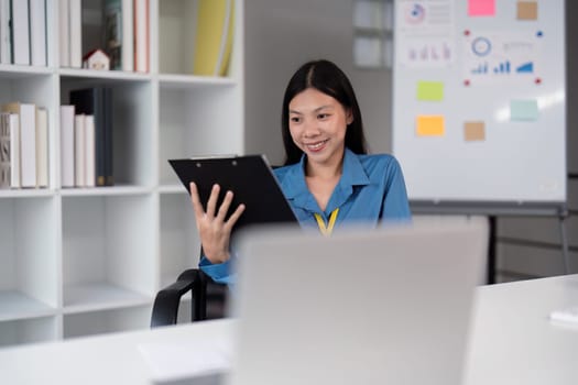 Businesswoman reviewing a clipboard while working on her laptop in a modern office, emphasizing professional analysis and productivity