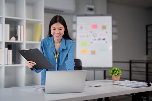 Successful businesswoman working on a laptop while holding a clipboard in a modern office, emphasizing professional achievement and productivity