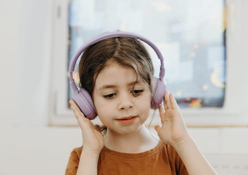 Portrait of a beautiful Caucasian brunette girl with big eyes sitting on a bed in a room by the window listening to her favorite music through headphones on a spring day, view from the side.