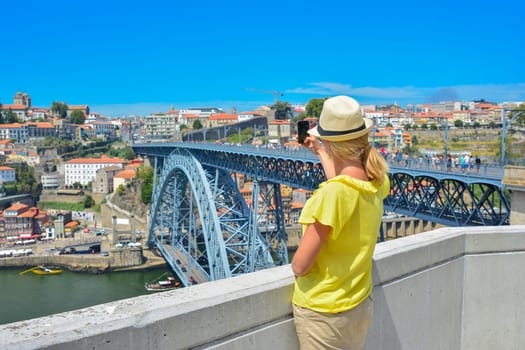 Blonde girl in a hat girl makes selfie on smartphone near Dom Luis Bridge in Porto, Portugal