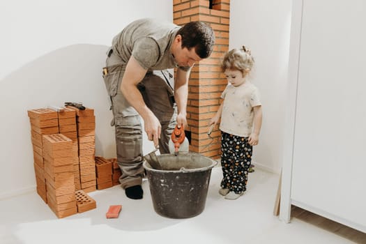 One young Caucasian man in a work uniform stands bent over and uses a spatula to knead cement mortar, and a little daughter stands nearby and carefully watches the process in the room where repairs and installation of a smoker for a fireplace made of bricks are underway, close-up side view. The concept is clean brickwork.
