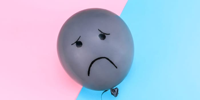 One gray balloon with black drawn sad eyes and a smile lies in the center on a blue-pink background with copy space on the sides, flat lay close-up.