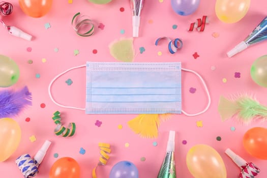 Many colored pastel inflated mini balloons with a medical mask, scattered confetti, tinsel, feathers and whistles lie in an oval frame on a pink background with copy space in the center, flat lay close-up.