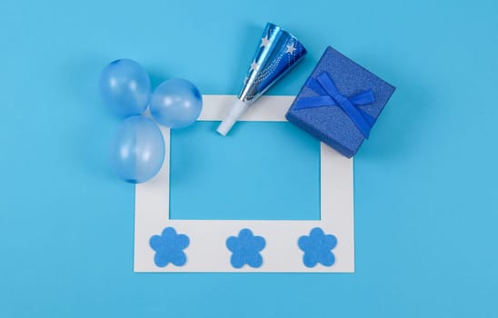 Empty paper frame, three inflated mini balloons, one whistle, decorative felt flowers and a shiny gift box lie on a blue background with copy space in the center, flat lay close-up.