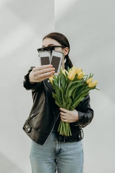 A young beautiful Caucasian brunette girl in sunglasses and a leather kurta with a bouquet of yellow tulips holds out her hand showing two packages of eyelashes for extensions standing near a white wall on a spring day, side view close-up with depth of field.