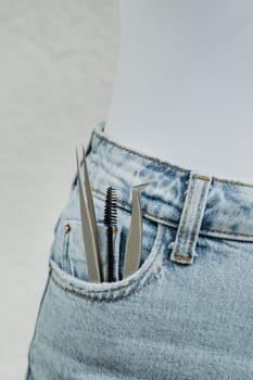 Two different tweezers and a round brush for eyelash extensions stick out from the right pocket of blue jeans, close-up side view.