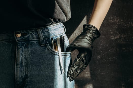 A set of brushes, tweezers and brushes for eyelash extensions stick out from the left pocket of blue jeans with a girl s hand in black gloves on a dark gray stone background, close-up side view. Dark style.