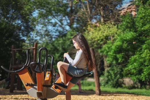Caucasian beautiful girl with long brown flowing hair, a gray long-sleeved t-shirt, shorts and glasses rides on a spinning swing in the park at the playground, close-up side view with selective focus. The concept of PARKS and REC, happy childhood, children's picnic, holidays, children's recreation, outdoors.