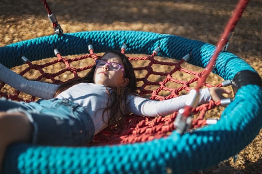Portrait of a beautiful caucasian girl of eight - nine years old in glasses, a long sleeve t-shirt and shorts with her eyes closed, rides lying on a round rope swing in a park on a playground, close-up side view. The concept of PARKS and SECs, happy childhood, children picnic, holidays, children recreation, outdoors, playgrounds, mental health.