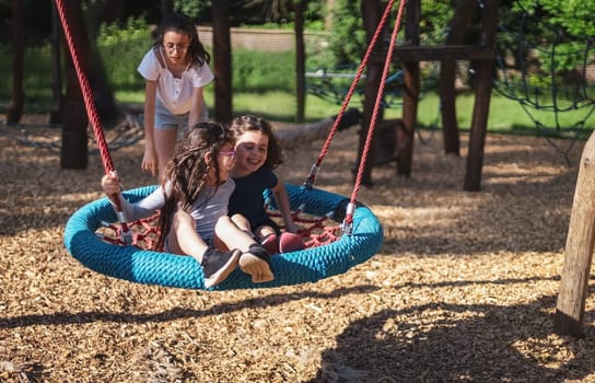 Portrait of a teenage girl swinging on a round rope swing with two laughing younger sisters in a park on a playground, close-up side view. The concept of PARKS and shopping centers, happy childhood, children's picnic, holidays, children's recreation, nature, playgrounds, mental health.