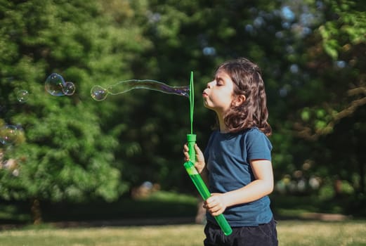 Portrait of one beautiful Caucasian brunette girl blowing a long soap bubble, standing on the right in the park on the playground, close-up side view. The concept of PARKS and RECREATION, happy childhood, children's picnic, happy childhood, outdoor recreation, playgrounds.