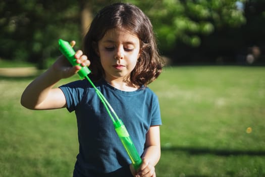 Portrait of a beautiful Caucasian brunette girl with a green bottle of soap bubbles standing in a park on a playground, close-up side view. The concept of PARKS and RECREATION, happy childhood, children's picnic, happy childhood, outdoor recreation, playgrounds.