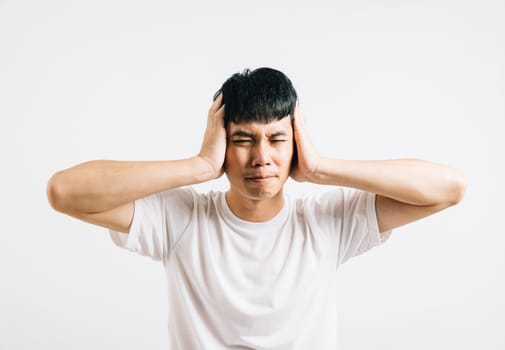 An Asian man in pain, using his hands to cover his ears and shutting his eyes to cope with the loud noise. Studio shot isolated on white background, showing his need for relief.