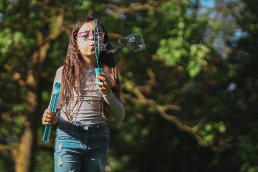 Portrait of one beautiful Caucasian brunette girl in glasses emotionally blowing bizarre shapes of soap bubbles, standing on the left in the park on the playground, close-up view from below. Concept of PARKS and RECREATION, happy childhood, children's picnic, happy childhood, outdoor recreation, playgrounds.