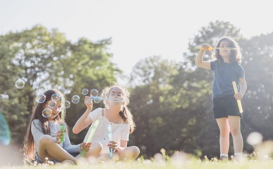 Portrait of three beautiful Caucasian children sisters blowing a lot of fancy soap bubbles, sitting in a park on the playground of a flower meadow under backlight, close-up view from below with selective focus. Concept PARKS and RECREATION, happy childhood, children's picnic, fairy-tale childhood, playgrounds, children's entertainment.