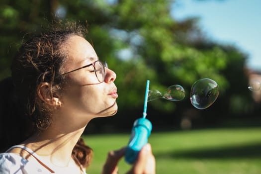 Portrait of one beautiful Caucasian teenage girl in glasses with closed eyes, blowing soap bubbles, standing sideways in a park on a playground, close-up side view. PARKS and RECREATION concept, happy childhood, children's picnic, happy childhood, outdoor recreation, playgrounds.