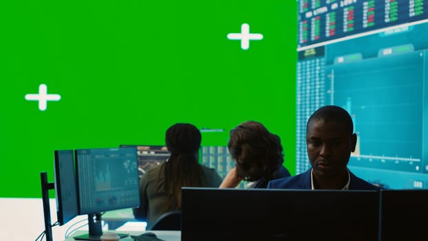 Diverse group of business specialists solve their daily tasks in control room with big greenscreen, analyzing big data numbers to ensure organizational progress. Startup management team. Camera A.