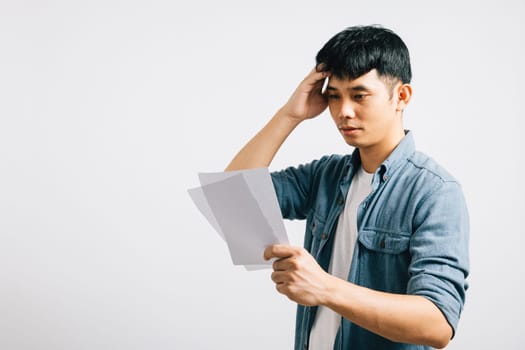 In a studio shot on white, a troubled Asian man holds a paper, calculating bills and displaying signs of financial strain, portraying his upset and stress over the situation. over bill