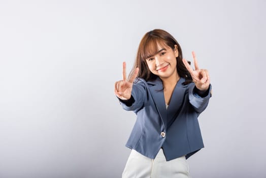 An Asian woman expresses her optimism and peace by forming victory and peace signs near her eye. This studio shot on a white background radiates positivity and success.