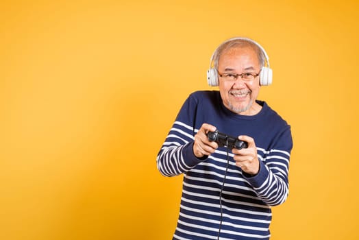 Portrait Asian smiling old man with headphones holding game controller studio shot isolated yellow background, senior man pensioner feeling winning playing a video game with joystick console