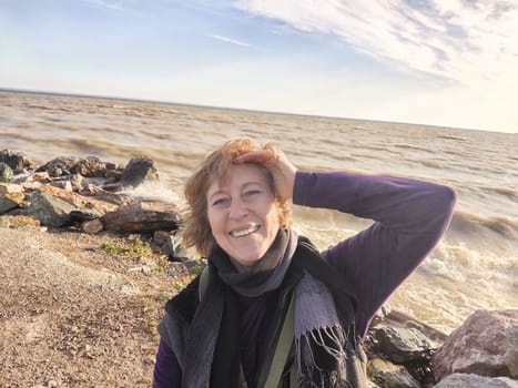 Happy cheerful middle aged woman in scarf taking selfie on nature outdoors and sun with water and waves of sea on the background