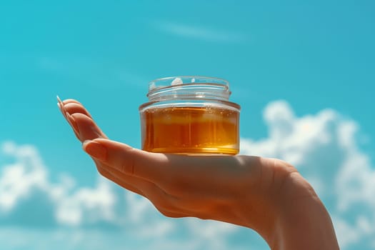 A hand holds a small glass jar of honey against a backdrop of a blue sky and white clouds.