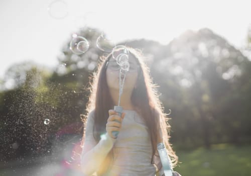 Blurred background of a beautiful Caucasian girl blowing soap bubbles in a meadow in a park on a playground in backlight, close-up side view with selective focus. The concept of PARKS and RECREATION, happy childhood, children's picnic, fairy-tale childhood, outdoor recreation, outdoors.