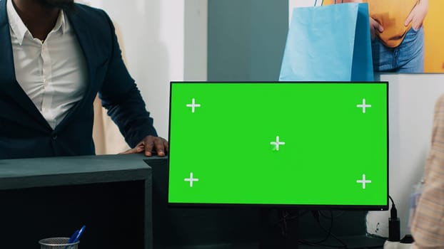 African american worker uses computer with greenscreen display on at checkout counter, putting clothes in a bag for customer. Employee looks at pc showing copyspace mockup screen. Camera A.