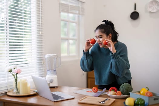 Young woman in a kitchen holding tomatoes, preparing vegetables on a cutting board, and video chatting via laptop, highlighting healthy cooking and online communication