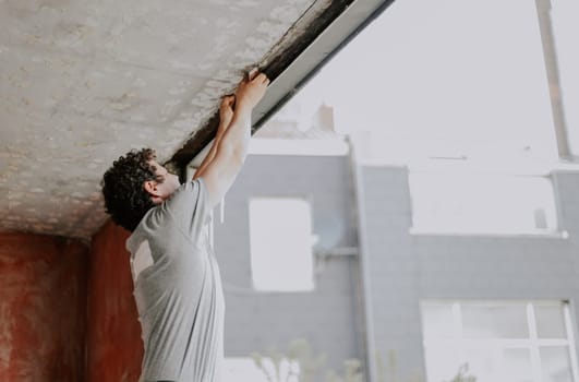 A young caucasian man in a gray t-shirt with curly brown hair is repairing a window opening, standing in a half turn and cleaning the frames with a hand with a construction knife, getting ready to install a window, close-up side view. The concept of home renovation, window installation, construction work, diy, lifestyle, at home, private services and small business.