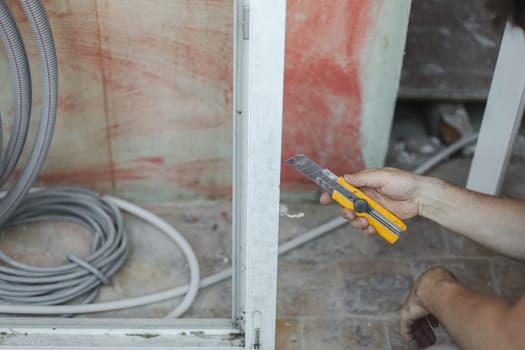 Young caucasian man holding a yellow construction knife on the background of a dirty frame near the wall, close-up side view. The concept of cleaning and installing windows, construction work, house renovation.