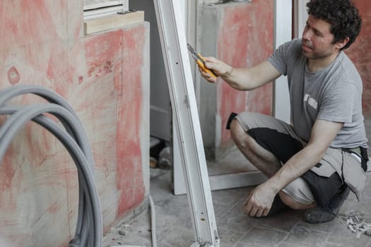 Young caucasian man holding a yellow construction knife and cleaning a dirty window frame while sitting on the floor against a wall, close-up side view. The concept of cleaning and installing windows, construction work, house renovation.