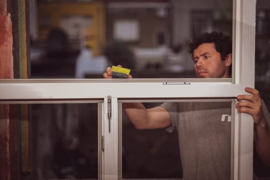 Handsome young caucasian satisfied man washing a window frame without glasses with a sponge and soap, preparing them for installation against a red wall, side view close-up. The concept of home renovation, installation of windows, construction work.