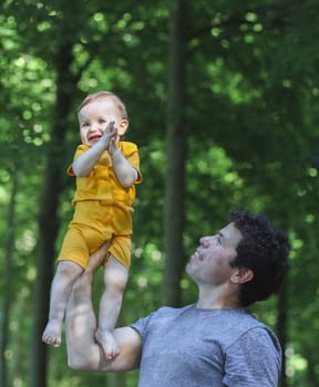 Caucasian young and beautiful male dad in a blue t-shirt holds his little daughter in a yellow bodysuit up high with his hand and cheerfully clapping her hands with happiness in a public park against the background of green trees, side view close-up. Concept of fatherhood, dads, family vacation, spring walk dad care.