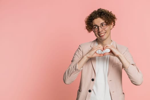 Man in love. Smiling attractive Caucasian young man with curly hair makes heart gesture demonstrates love sign expresses good positive feelings and sympathy. Guy isolated on pink background