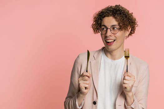 Ready to eat. Hungry Caucasian man with curly hair waiting for serving dinner dishes with with restlessness holding cutlery fork knife, will appreciate delicious restaurant meal on pink background