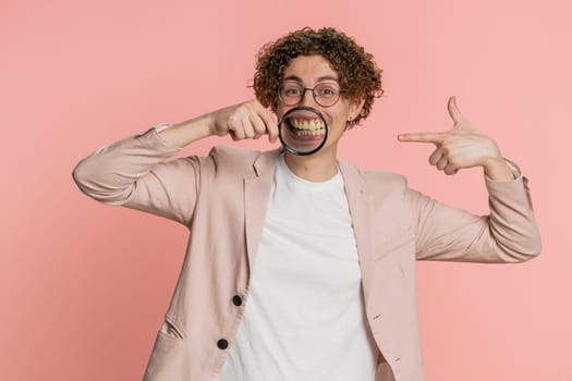 Happy Caucasian man holding magnifier glass on teeth, looking at camera, showing funny smiling silly face mouth. Health care, hygiene, stomatology. Curly haired young guy isolated on pink background