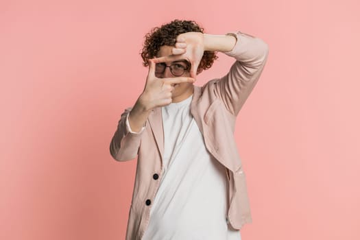 Caucasian man photographer gesturing picture frame with hands, looks through fingers and focusing on interesting moment, imitating zoom and cropping nice image. Young guy isolated on pink background