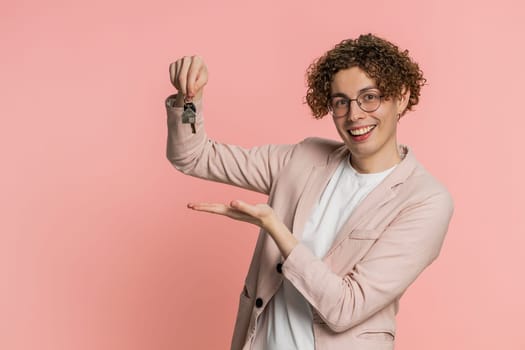 Caucasian young man with curly hair real estate agent lifting hand showing the keys of new home house apartment, buying or renting property, mortgage loan, insurance. Guy isolated on pink background