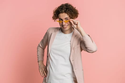 Stylish smiling Caucasian young man with curly hair wearing orange sunglasses glad expression looking at camera dreaming, resting relaxation feel satisfied good news. Guy isolated on pink background