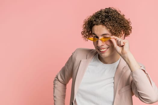 Stylish smiling Caucasian young man with curly hair wearing orange sunglasses glad expression looking at camera dreaming, resting relaxation feel satisfied good news. Guy isolated on pink background