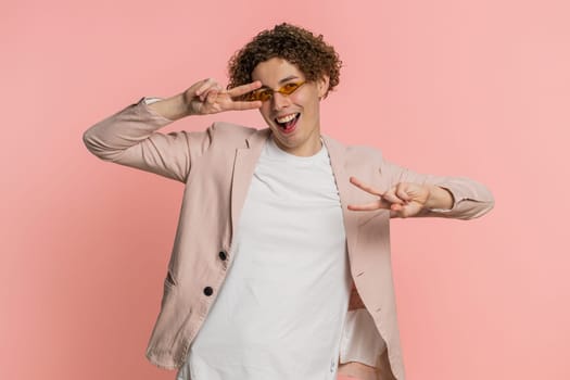 Caucasian young man with curly hair listening music, dancing disco fooling around having fun expressive gesticulating hands. Music app advertisement. Happy guy isolated on studio pink background