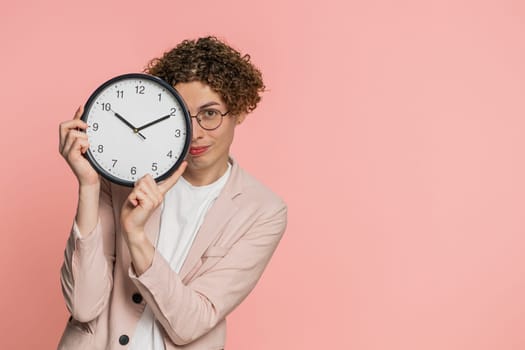 Happy smiling Caucasian curly haired man in jacket holding office clock hiding behind, checking time on watch, running late to work, being in delay, deadline obscuring face on pink background indoors