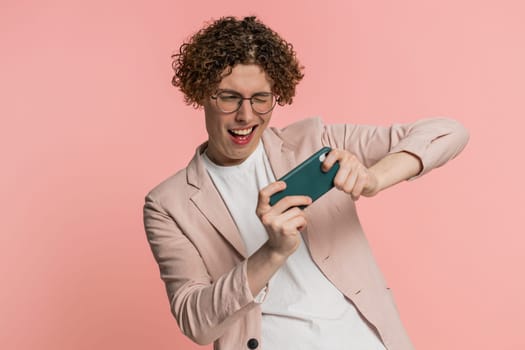 Happy excited Caucasian young man with curly hair playing racing drive simulator shooter video games on smartphone. Guy player celebrate victory lottery jackpot goal achievement on pink background