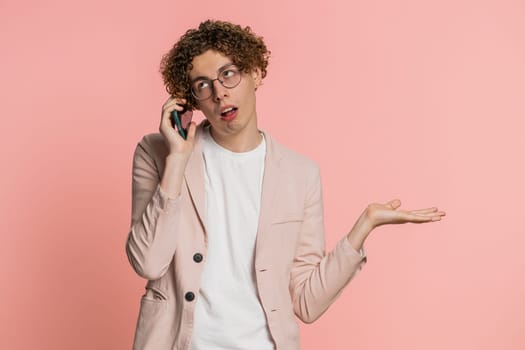 Disinterested man with curly hair having annoyed boring talk on smartphone. Guy tired sleepy freelancer talking on mobile phone with friend, making online conversation isolated on pink background