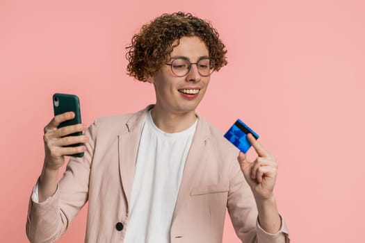 Happy Caucasian young man with curly hair using credit bank card and smartphone while transferring spending money, purchases online shopping, ordering food delivery. Guy isolated on pink background